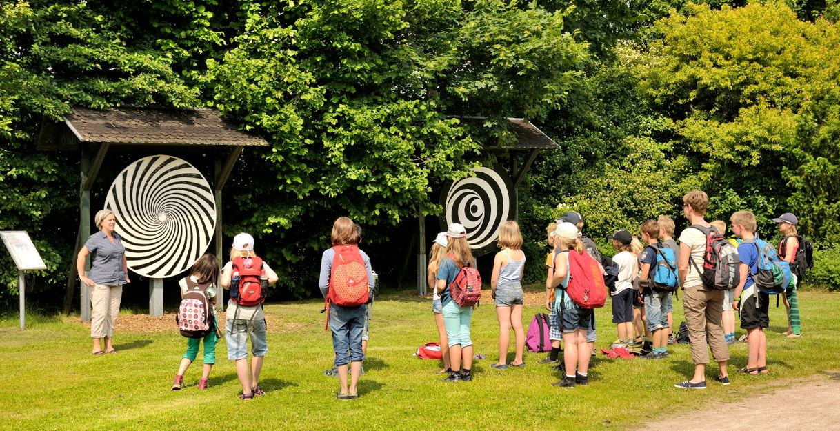 Eine Gruppe von Kindern mit Rucksäcken steht vor einer spiralförmigen Kunstinstallation im Freien, umgeben von Bäumen.
