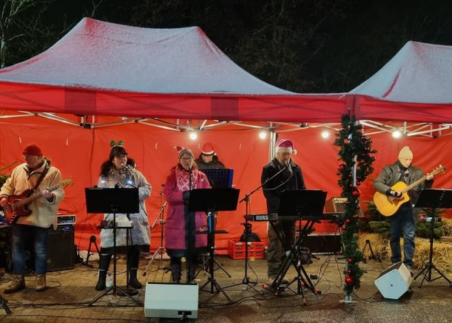Eine Band spielt unter einem roten Pavillon bei einem Weihnachtsmarkt. Die Mitglieder tragen winterliche Kleidung und Weihnachtsmützen.