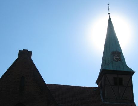 Kirchturm mit Uhr vor strahlend blauem Himmel und Sonne im Hintergrund.