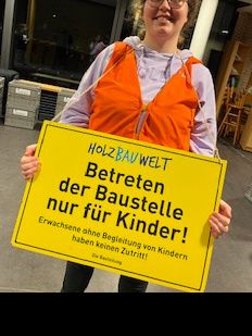Eine Person hält ein gelbes Schild mit der Aufschrift: 'Betreten der Baustelle nur für Kinder! Erwachsene ohne Begleitung von Kindern haben keinen Zutritt!'