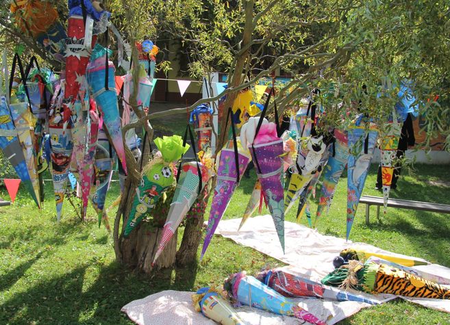 Ein Baum, an dem bunte Schultüten hängen, steht auf einer Wiese. Einige Schultüten liegen auf Decken am Boden.