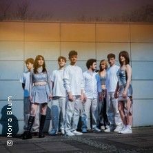 Eine Gruppe von acht jungen Erwachsenen steht vor einer modernen, grauen Wand. Sie tragen modische, überwiegend graue und weiße Kleidung.