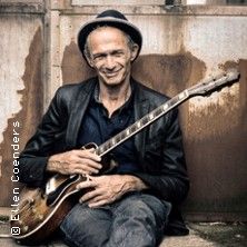 Ein Mann mit Hut und Gitarre sitzt lächelnd vor einer rostigen Wand.