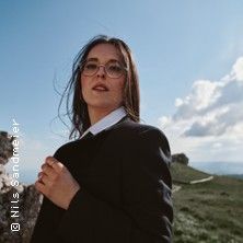 Eine Frau mit Brille und dunklem Blazer steht in einer grünen Landschaft unter einem blauen Himmel.