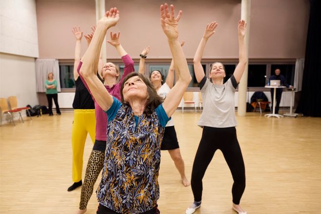 Fünf Personen stehen in einem Tanzstudio hintereinander und haben je beide Arme in Richtung Decke gestreckt. Sie tragen bunte Sportkleidung und ihre Blicke sind in Richtung Decke gerichtet.