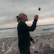 Eine Person steht am Strand und wirft einen Stein in die Luft. Der Himmel ist bewölkt und das Meer ist im Hintergrund zu sehen.