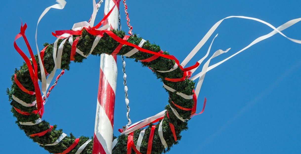 Ein geschmückter Maibaum mit Kranz und roten sowie weißen Bändern vor blauem Himmel.