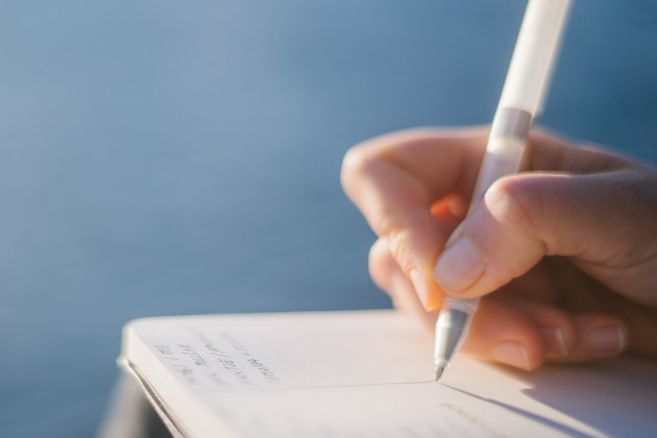 Nahaufnahme einer Hand, die mit einem Stift in ein Notizbuch schreibt, vor einem verschwommenen blauen Hintergrund.