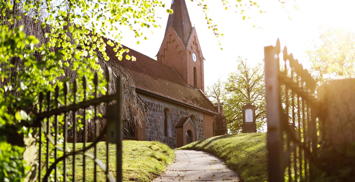 Ein Weg führt durch ein geöffnetes Tor zu einer alten Kirche mit rotem Backsteinturm, umgeben von Bäumen im Sonnenlicht.
