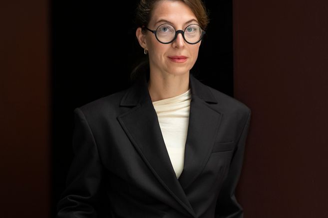 Eine Frau mit Brille und schwarzem Blazer steht vor einem dunklen Hintergrund.