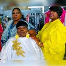Drei Frauen in eleganter Kleidung posieren in einem Raum mit Kleidung im Hintergrund.