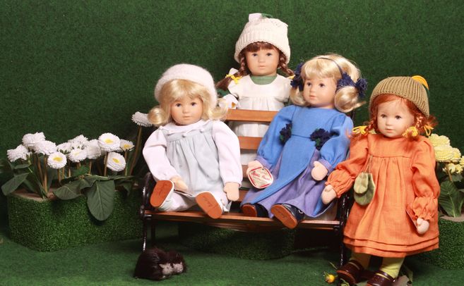 Vier Puppen in bunter Kleidung sitzen auf einer Bank vor einem grünen Hintergrund mit Blumen.