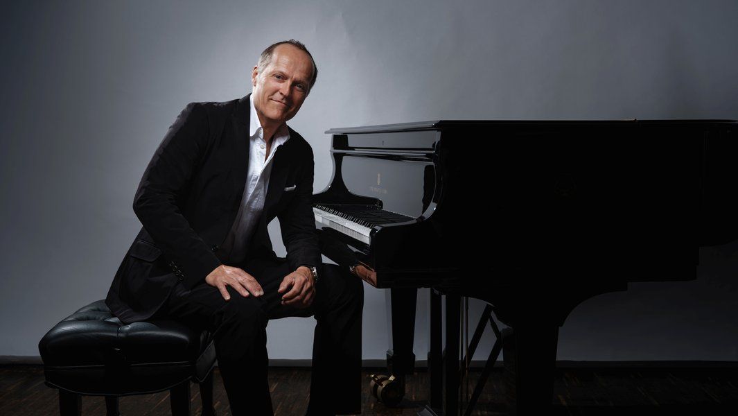 Ein Mann in einem Anzug sitzt neben einem Flügel auf einem Hocker.