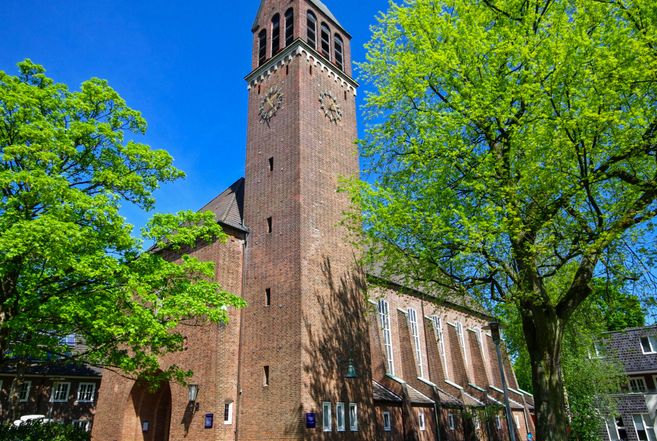 Eine Backsteinkirche mit einem hohen Turm und einer Uhr, umgeben von grünen Bäumen unter einem klaren blauen Himmel.