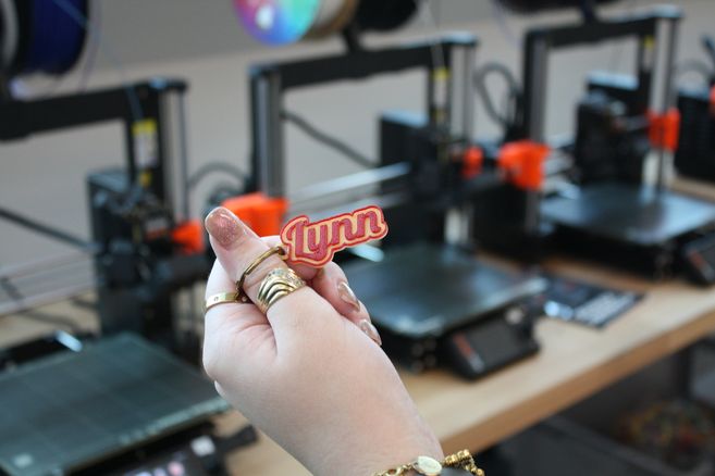 Hand hält einen kleinen 3D-gedruckten Anhänger mit der Aufschrift „Lynn“ vor mehreren 3D-Druckern.