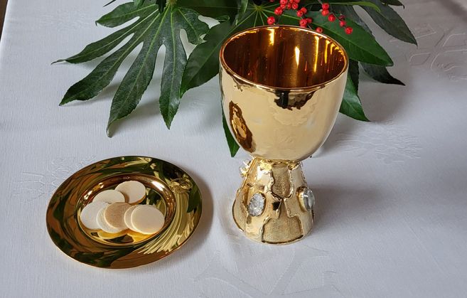 Ein goldener Kelch und eine Patene mit Hostien auf einem weißen Tuch, dekoriert mit grünen Blättern und roten Beeren.
