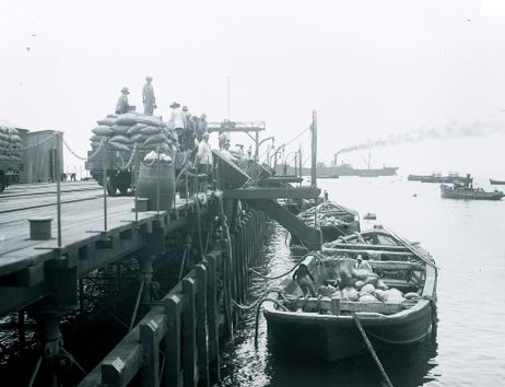 Arbeiter verladen Säcke auf einem Pier, Boote im Wasser, Dampfschiff im Hintergrund.