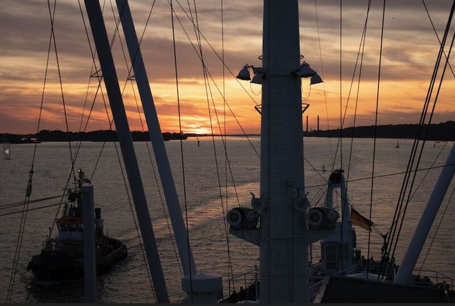 Sonnenuntergang über einem Fluss mit Schiffsmasten im Vordergrund.