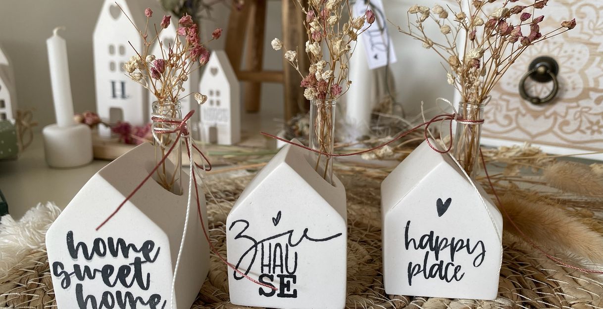 Drei kleine weiße Häuser aus Keramik mit Aufschriften und Trockenblumen.