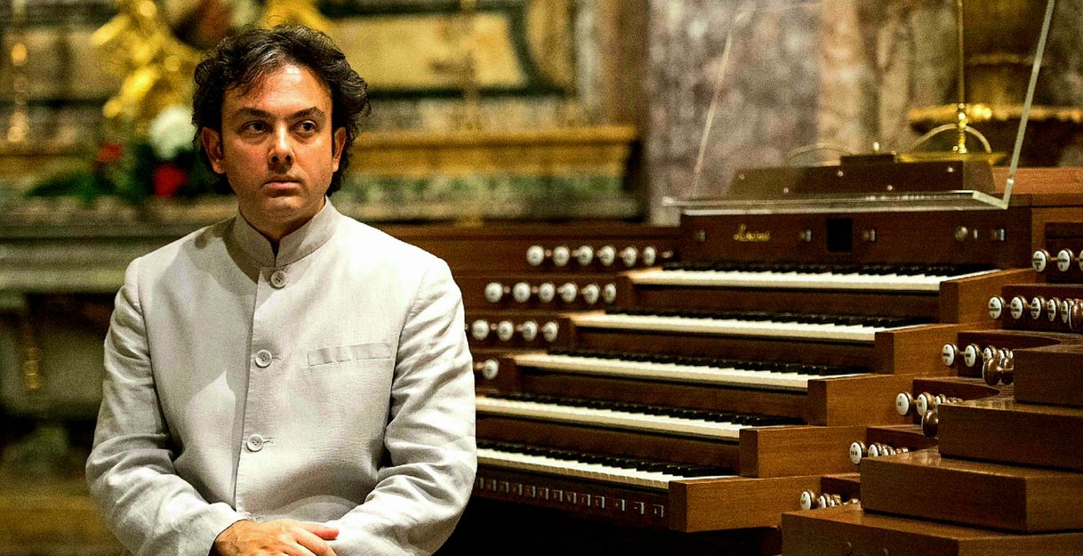 Ein Mann in einem hellen Anzug sitzt neben einer großen Orgel in einer Kirche.