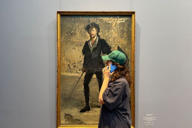 Foto eines Menschen mit langen lockigen Haaren, weitem Hemd und Kappe vor einem Gemälde. Die Person hält ein Handy an das Ohr während sie sich das Gemälde anschaut.