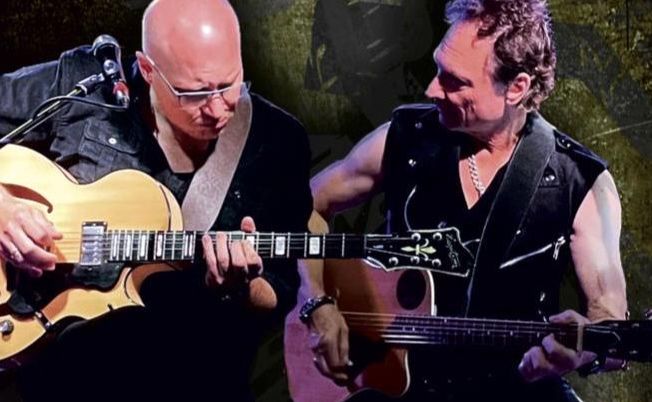 Zwei Männer spielen Gitarren auf einer Bühne.