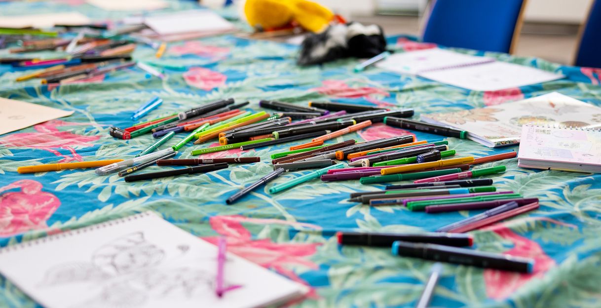Ein Tisch mit vielen bunten Stiften und Malbüchern auf einer tropisch gemusterten Tischdecke.
