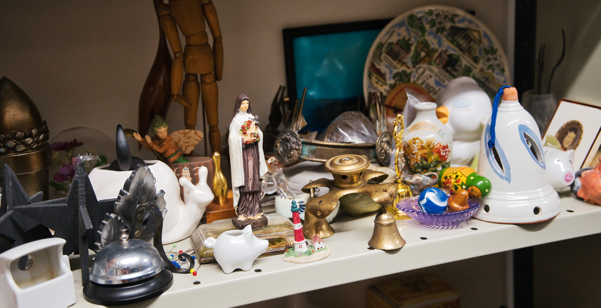 Der Requisitenfundus im SchauSpielHaus. Ein Regel mit diversen Requisiten, darunter ein Schwein aus Porzellan, eine kleine goldene Glocke, ein Männchen aus Holz, eine Marien-Statue und weitere Requisiten.