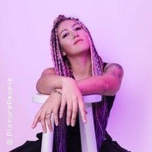 Frau mit bunten Zöpfen sitzt auf einem Hocker vor einem lila Hintergrund.