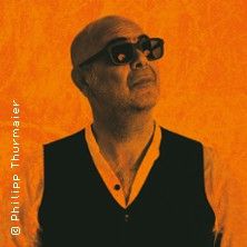 Ein Mann mit Glatze und Sonnenbrille schaut nach oben. Er trägt ein Hemd und eine Weste. Der Hintergrund ist orange.