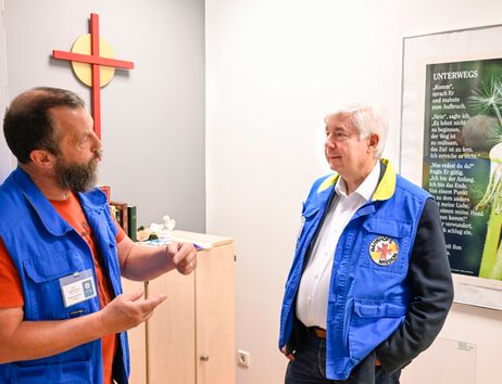 Zwei Männer in blauen Westen der Bahnhofsmission Bremen stehen in einem Raum mit Kreuz und Poster und unterhalten sich.