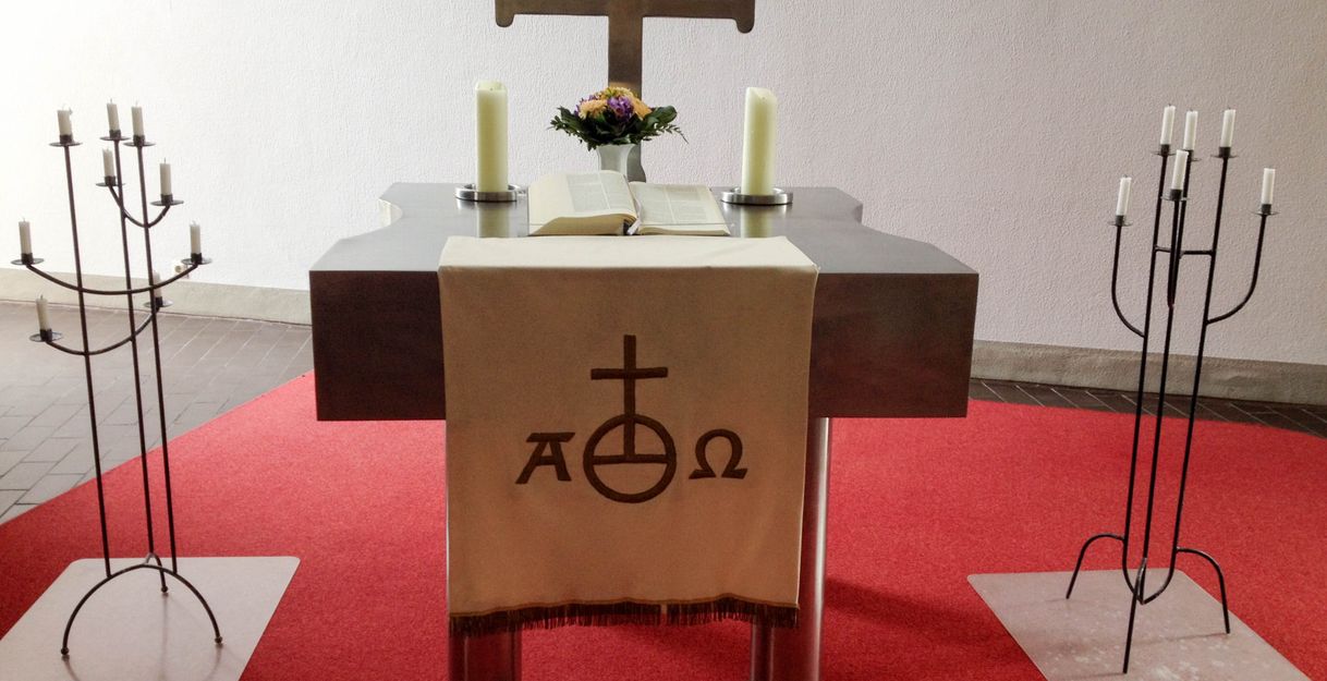 Ein Kirchenaltar mit einem Kreuz, zwei Kerzen und einem aufgeschlagenen Buch auf einem roten Teppich.