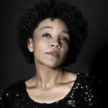 Porträt einer Frau mit lockigem Haar und schwarzem Paillettenoberteil vor dunklem Hintergrund.