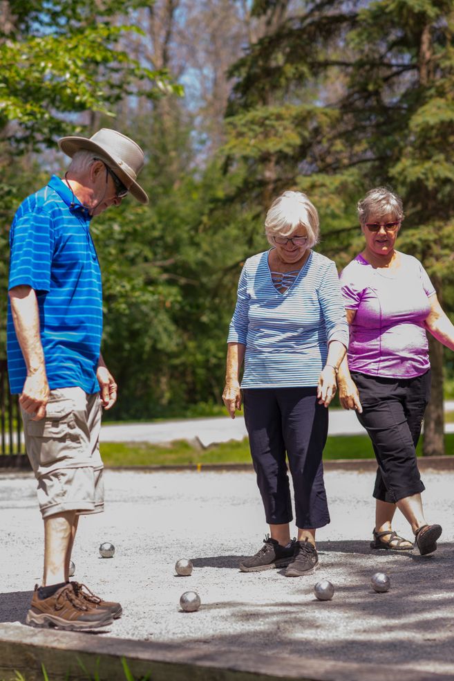 Drei ältere Personen spielen Boule im Freien auf einem Kiesplatz.