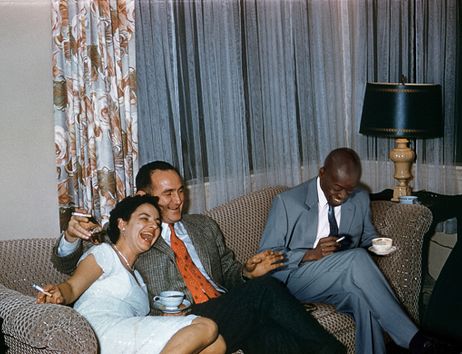 Drei Personen sitzen lachend auf einem Sofa, zwei halten Tassen, eine Person raucht. Im Hintergrund sind Vorhänge und eine Lampe zu sehen.