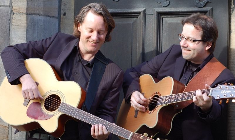 Zwei Männer spielen Akustikgitarren vor einer Tür.