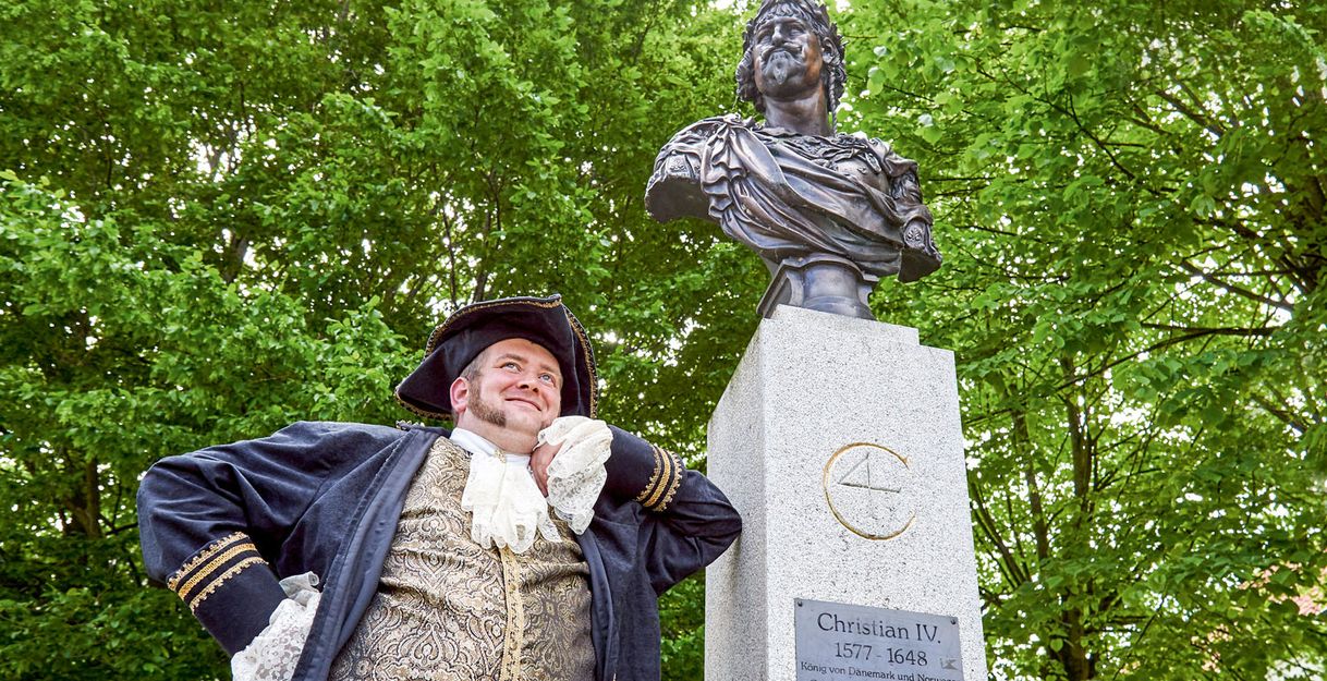 Ein Mann in historischer Kleidung steht neben einer Büste von Christian IV. vor einem Hintergrund aus grünen Bäumen.