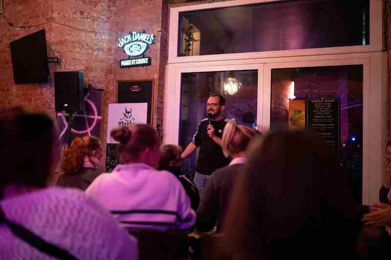 Ein Comedian steht mit Mikrofon vor einem Publikum in einer Bar.