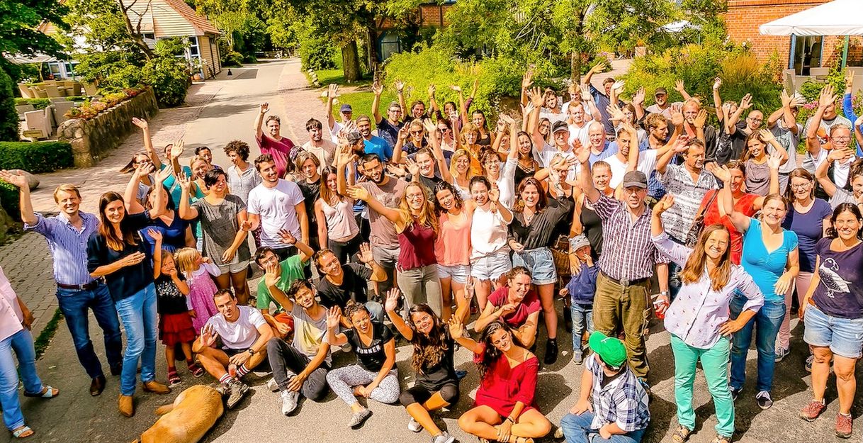 Eine große Gruppe von Menschen versammelt sich im Freien und winkt fröhlich in die Kamera.