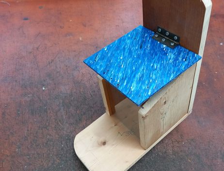 Fertig gebautes Futterhaus aus Holz mit einem blauen Dach aus recycelten Plastik mit Scharnier und einer Scheibe um Nüsse von Außen zu sehen. Das Haus hat davor eine Fläche wo das Eichhörnchen sitzen kann.