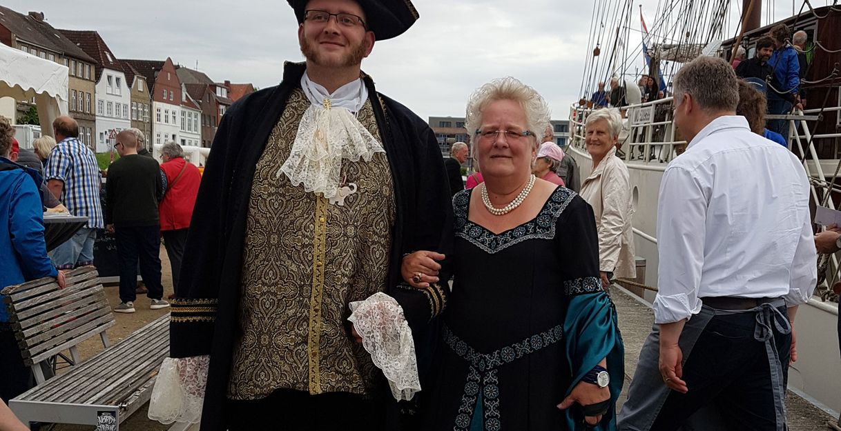 Ein Mann und eine Frau in historischen Kostümen stehen auf einem Hafenfest vor einem Segelschiff.