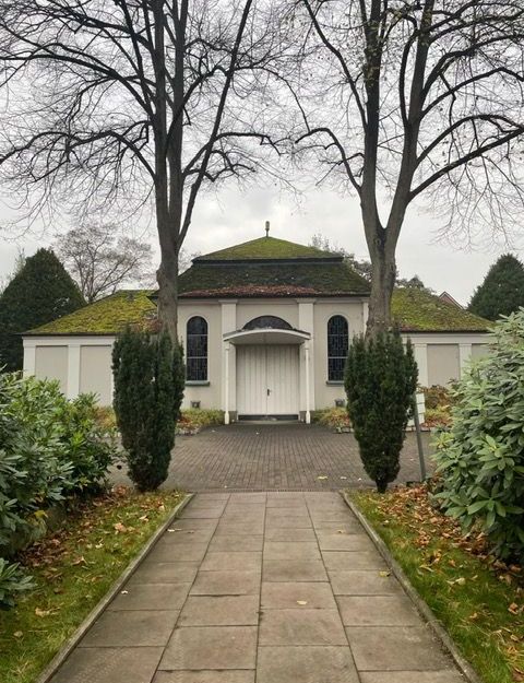 Eine kleine Friedhofskapelle mit moosbewachsenem Dach, flankiert von zwei Bäumen, umgeben von grünen Büschen und einem gepflasterten Weg.