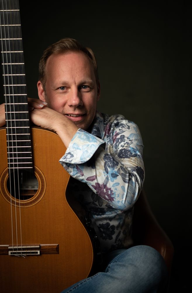 Ein Mann lehnt sich an eine klassische Gitarre und lächelt in die Kamera.