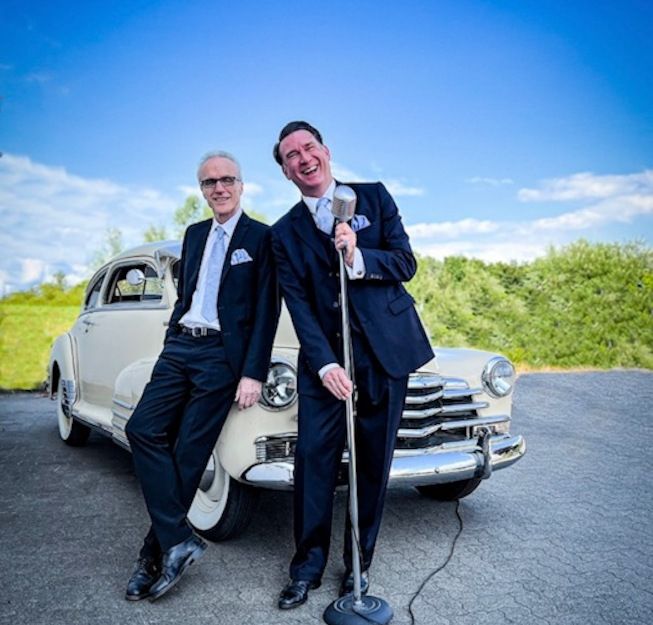 Zwei Männer in Anzügen lehnen an einem Oldtimer, einer hält ein Mikrofon. Der Himmel ist blau und klar.