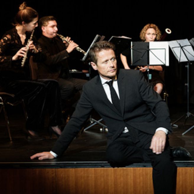 Ein Mann in einem Anzug sitzt auf einer Bühne, während im Hintergrund Musiker mit Blasinstrumenten spielen.