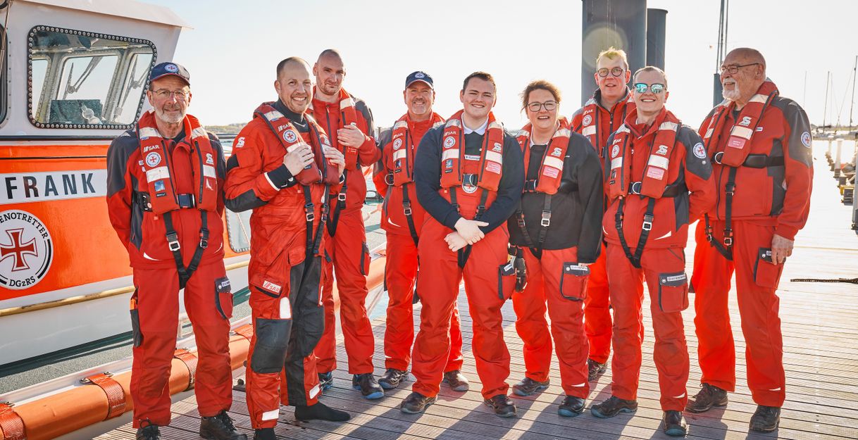 Eine Gruppe von Menschen in roten Rettungsanzügen steht auf einem Steg vor einem Rettungsboot.