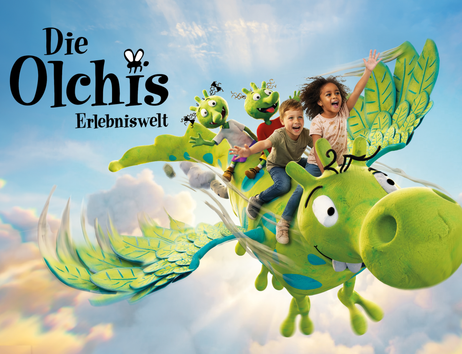 Zwei Kinder fliegen gemeinsam mit zwei grünen Olchi-Figuren auf dem großen grünen Drachen Feuerstuhl durch einen blauen Himmel voller Wolken.