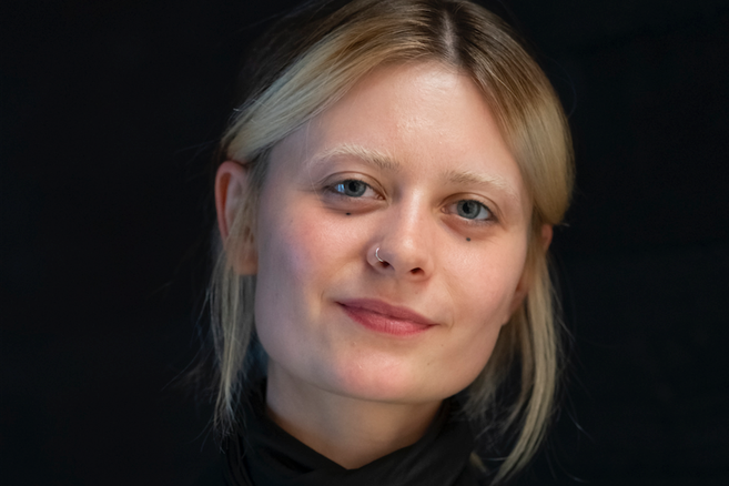 Porträt einer jungen Frau mit blonden Haaren und Nasenpiercing vor schwarzem Hintergrund.