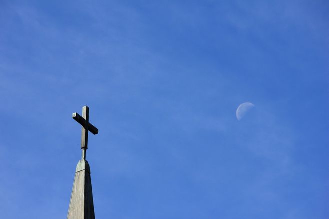 Ein Kirchturm mit einem Kreuz vor einem blauen Himmel und einem halb sichtbaren Mond.