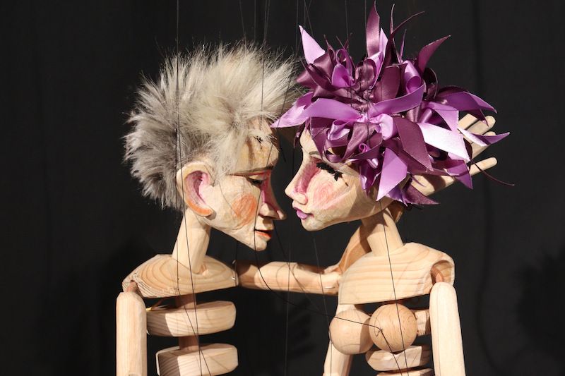 Zwei Holzmarionetten, eine mit lila Bändern im Haar, stehen sich nah gegenüber auf schwarzem Hintergrund.